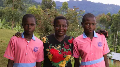 Retrato de Rebecca Nakayega Wandegge. De pie en una zona rural, sonríe junto a sus dos hijas que llevan el uniforme escolar. (Uganda, 2025)